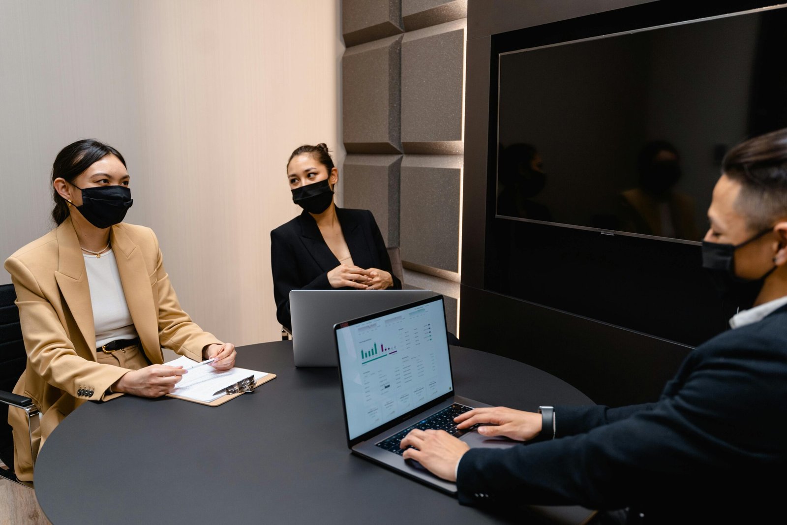 Business team in masks discussing around a table with laptops showing data.